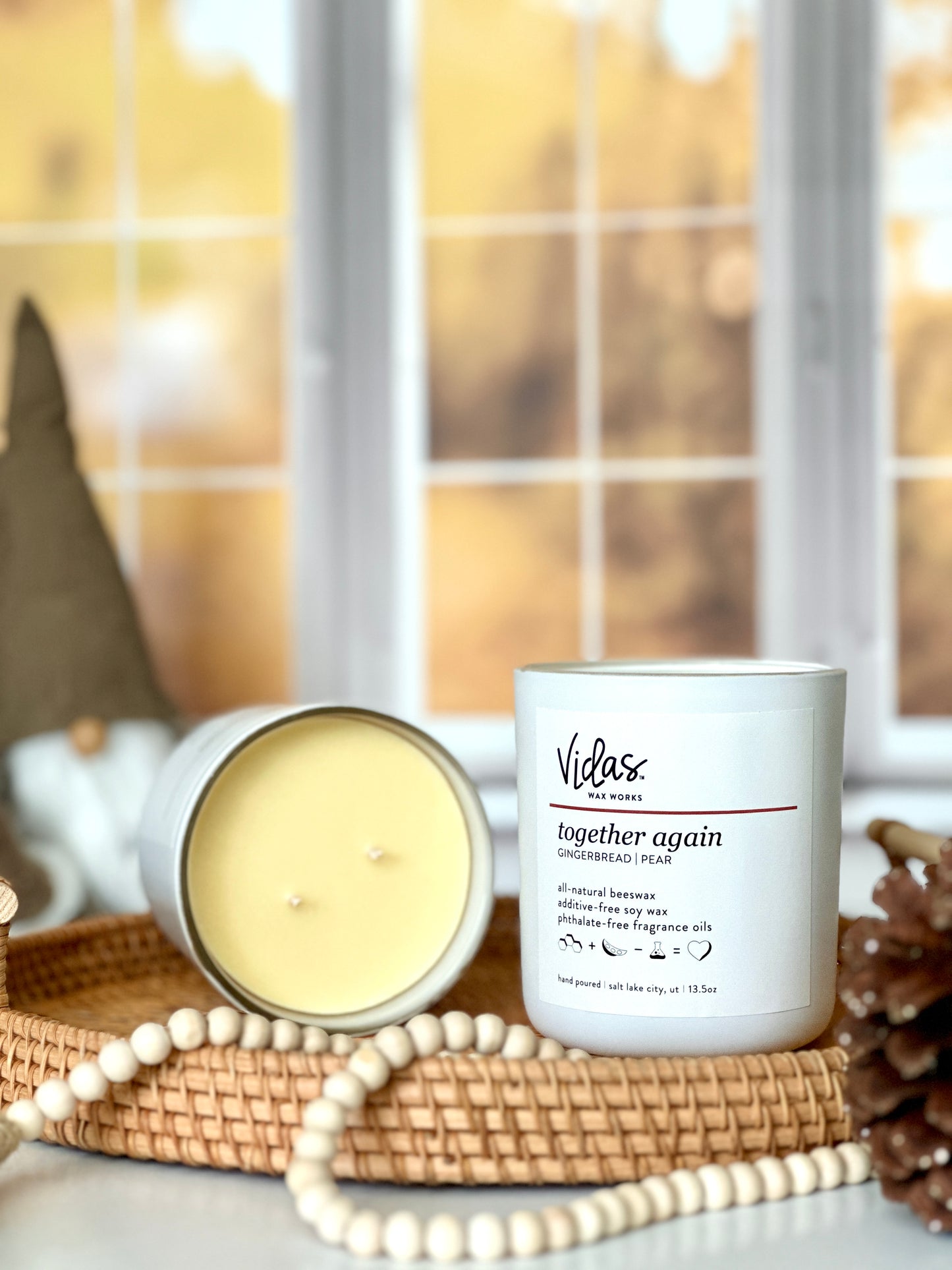 Captivating fall fragrance: Gingerbread and pear. An open 13.5oz travel candle reveals its natural blend of beeswax and soy wax on a wicker serving tray, next to another candle. A string of beads and a pinecone grace the foreground, while a whimsical brown gnome adds charm against the backdrop of blurred yellow fall leaves outside a window.