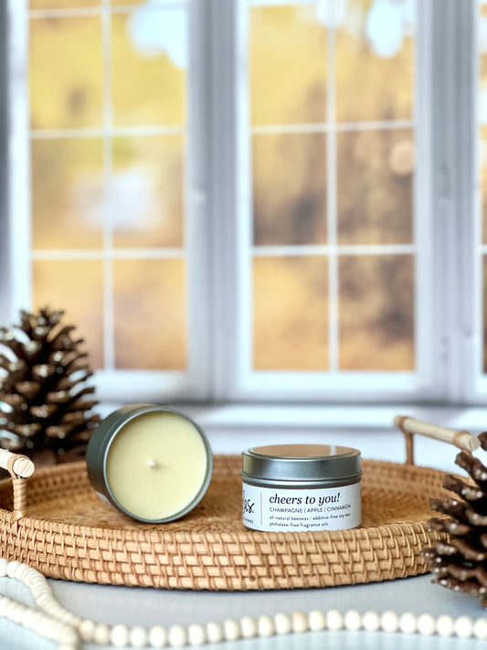 Indulge in the scent of fall: Champagne, apple, and cinnamon. A wicker serving tray cradles two 3.5oz candle travel tin, while pinecones and a string of decorative beads add rustic charm. A backdrop of blurred yellow fall leaves outside a window brings the season's ambiance to life.