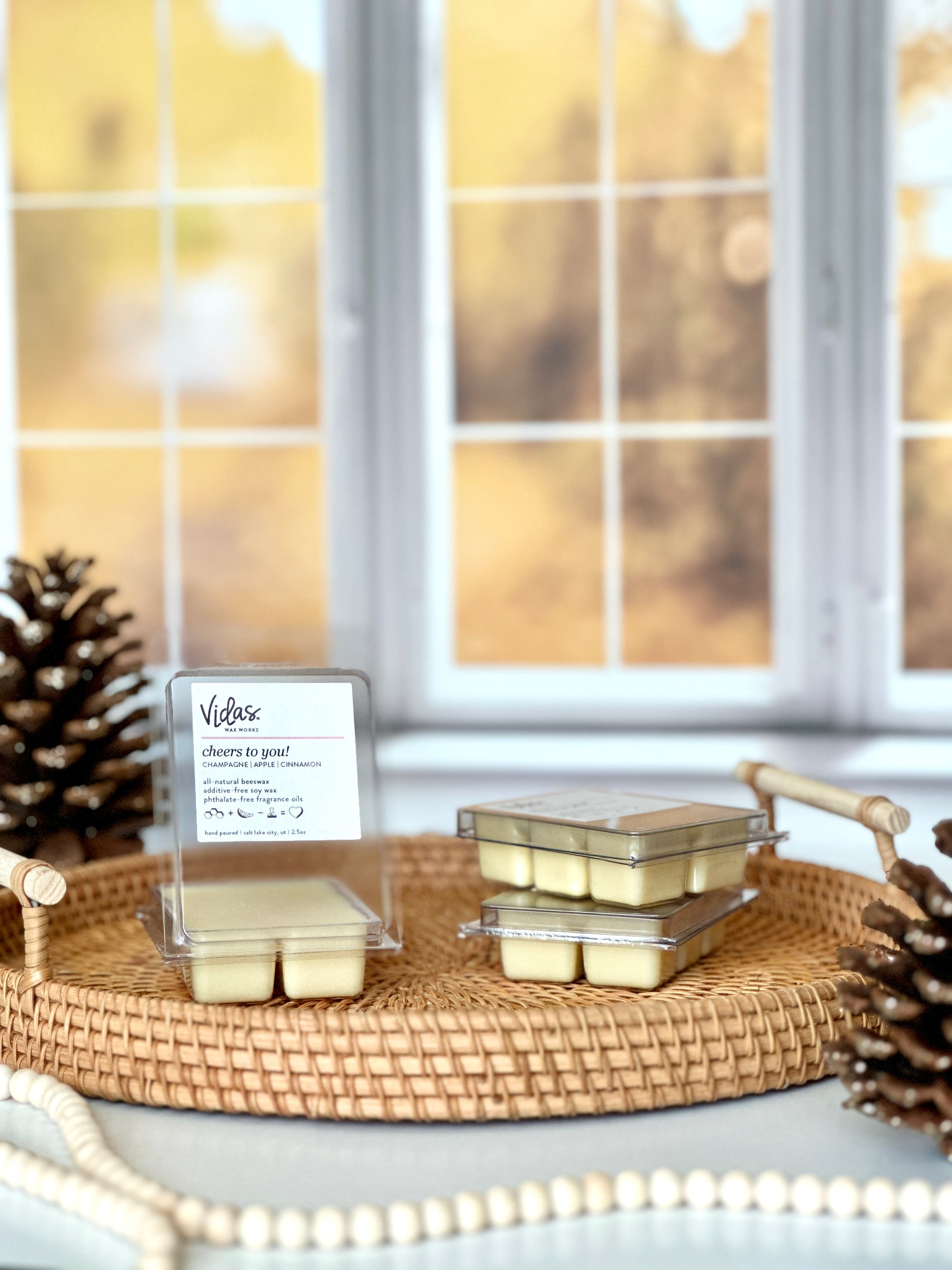 Indulge in the scent of fall: Champagne, apple, and cinnamon. A wicker serving tray cradles an open 2.5oz clamshell of wax melts, while pinecones and a string of decorative beads add rustic charm. A backdrop of blurred yellow fall leaves outside a window brings the season's ambiance to life.