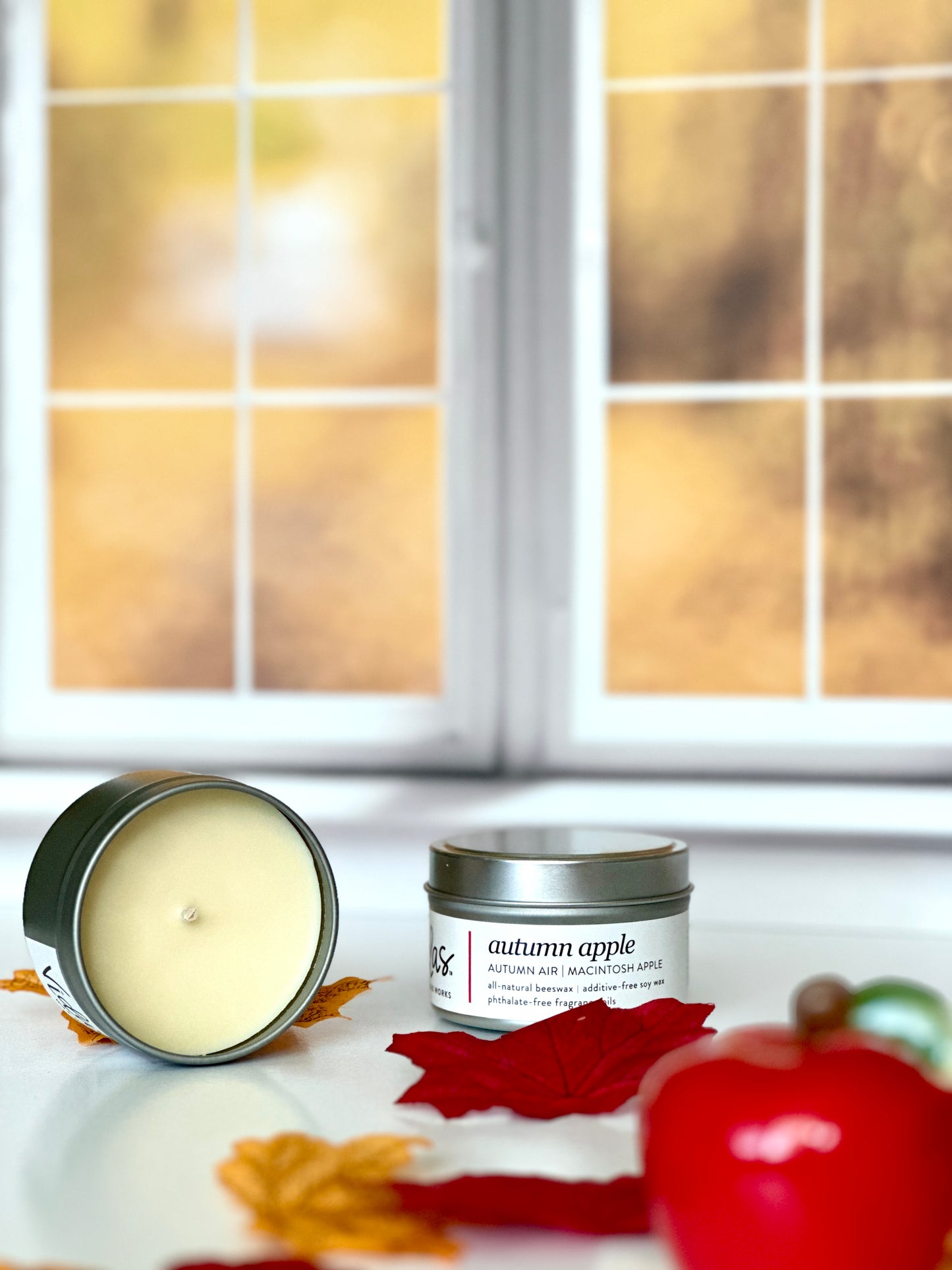 Captivating fall fragrance: Autumn air and Macintosh apple candle travel tin. A vivid red apple stands prominently in the foreground, surrounded by fall-colored leaves. Beyond, a blurred vista of yellow fall foliage outside a window sets the scene, capturing the essence of the season.