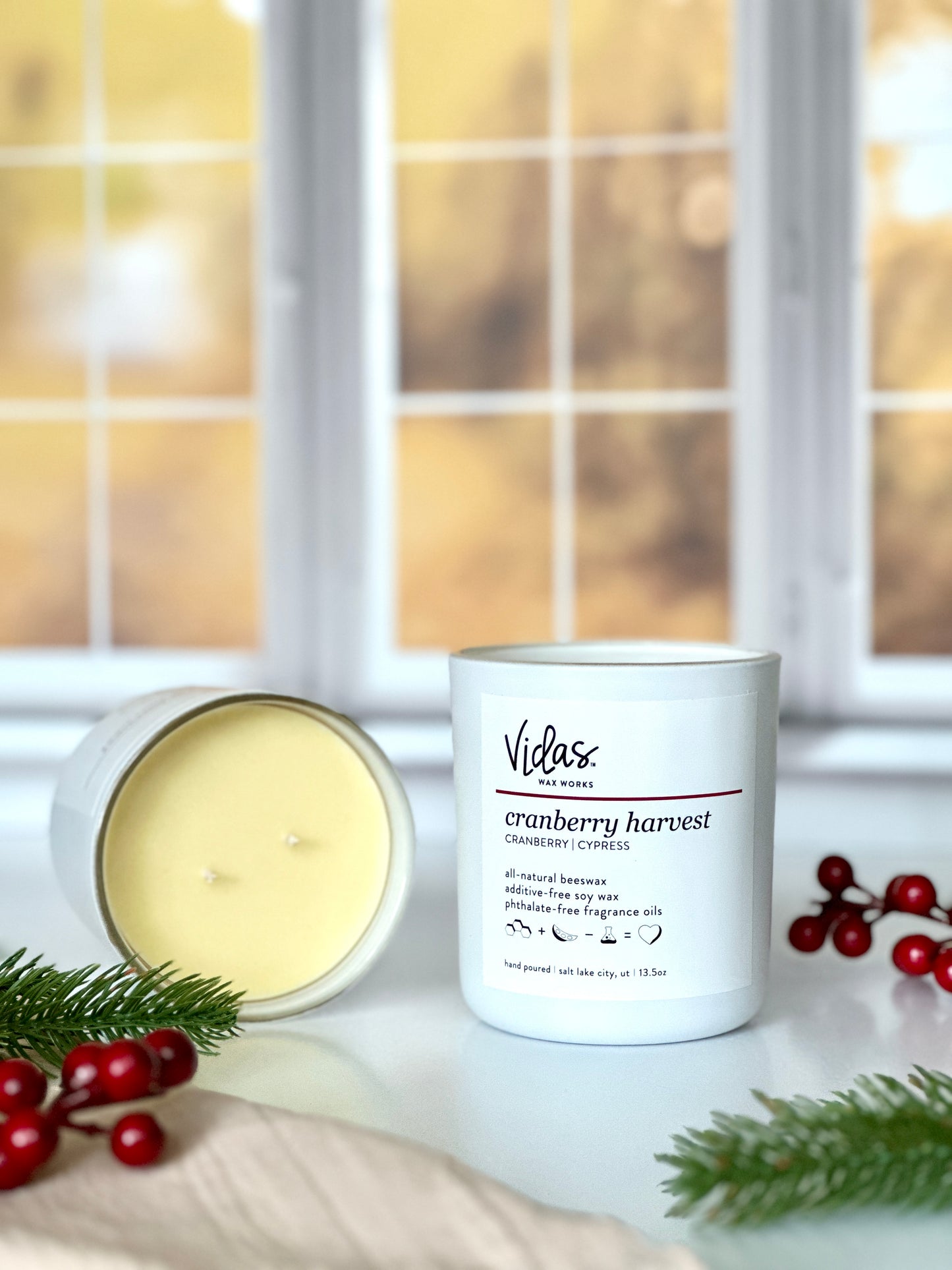 Embrace the essence of fall with our cranberry and cypress fragrance. A 13.5oz candle lays open, showcasing its beautiful yellow tinted beeswax and soy blend beside another candle. Cranberry strands frame the scene, while cypress adds a touch of nature's beauty. The backdrop of blurred yellow fall leaves outside a window completes the seasonal display.