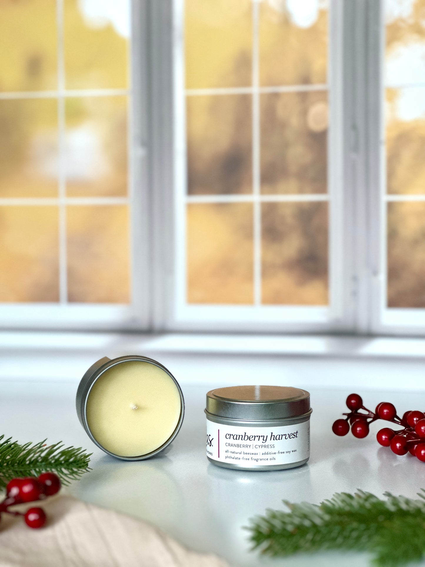 Embrace the essence of fall with our cranberry and cypress fragrance. A 3.5oz candle travel tin lays open, showcasing its beautiful yellow tinted beeswax and soy blend beside another travel tin. Cranberry strands frame the scene, while cypress adds a touch of nature's beauty. The backdrop of blurred yellow fall leaves outside a window completes the seasonal display.