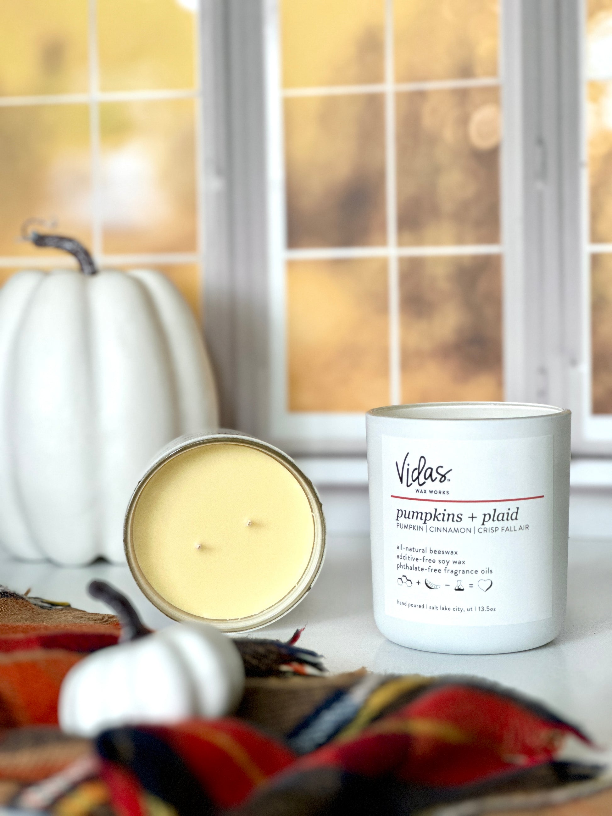 Capturing fall's allure: Pumpkins, cinnamon, and crisp fall air scent. A 13.5oz candle lays open showcasing its yellow tinted all-natural beeswax soy wax blend color next to another candle. A plaid scarf adorned with a small white pumpkin graces the foreground, while a taller white pumpkin rests against the backdrop of blurred yellow fall leaves outside a window, echoing the season's charm.