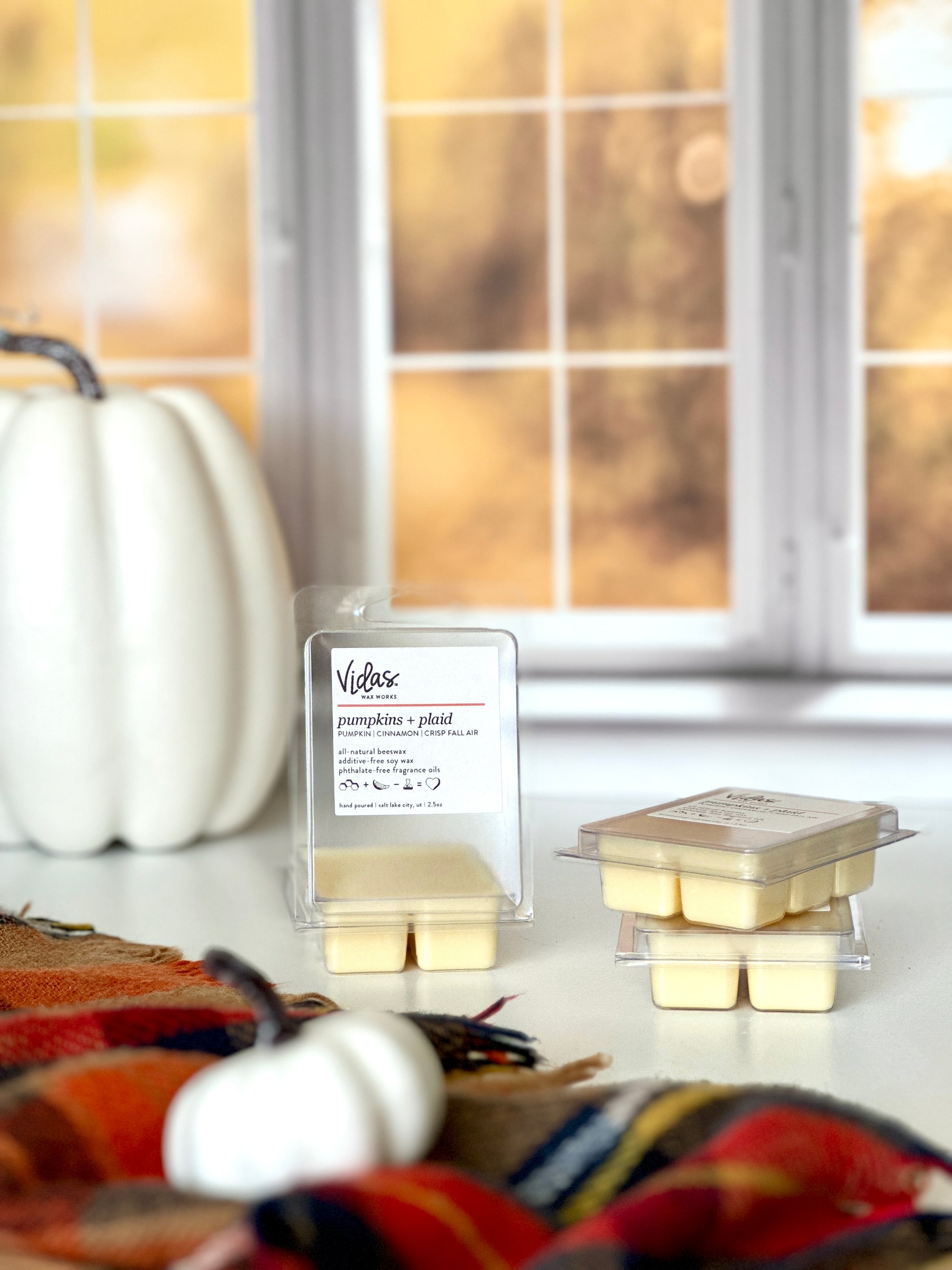 Capturing fall's allure: Pumpkins, cinnamon, and crisp fall air scent. A 2.5oz clamshell of wax melts sits open beside two neatly stacked packages. A plaid scarf adorned with a small white pumpkin graces the foreground, while a taller white pumpkin rests against the backdrop of blurred yellow fall leaves outside a window, echoing the season's charm.