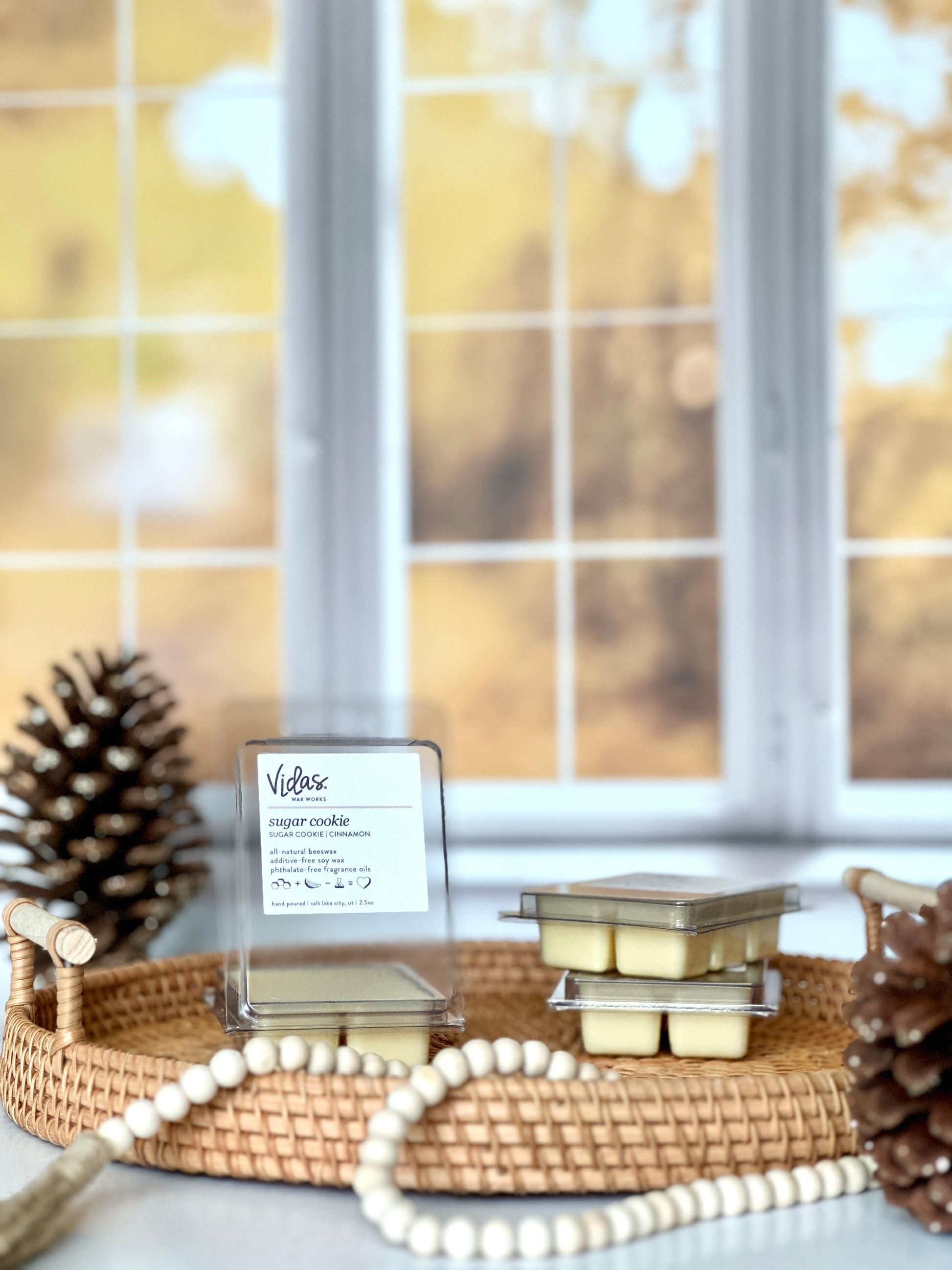 Experience the essence of fall: Sugar cookie and cinnamon fragrance. An open 2.5oz clamshell of wax melts rests alongside two stacked packages on a wicker serving tray. A pinecone graces the foreground, adorned with a string of beads, while a larger pinecone punctuates the scene against the backdrop of blurred yellow fall leaves outside a window.