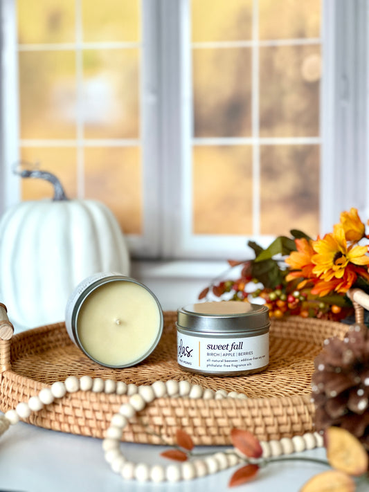 Captivating fall fragrance: Birch, apple, and berries. An open 3.5oz travel candle tin reveals its natural beeswax and soy wax blend on a wicker serving tray, accompanied by another tin. A pinecone and string of beads grace the foreground, while fall-colored flowers and a large pumpkin create a vibrant scene against the blurred backdrop of yellow fall leaves outside a window.