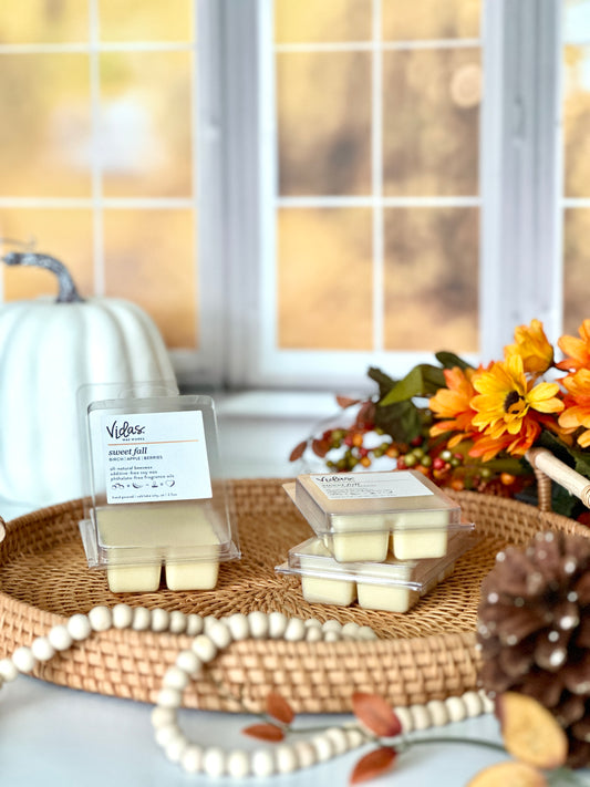 Embrace autumn's allure: birch, apple, and berries fragrance. An open 2.5oz clamshell sits on top of a wicker serving tray beside two neatly stacked packages. A pinecone and string of beads adorn the foreground, while fall-colored flowers and a large pumpkin create a vibrant backdrop against the blurred yellow fall leaves outside a window.