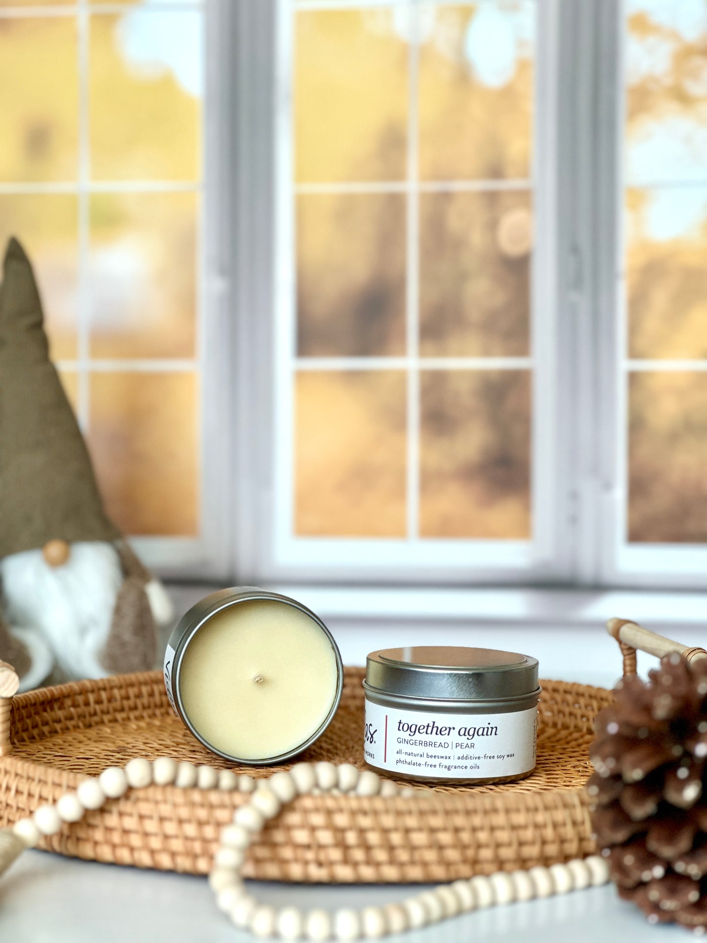 Captivating fall fragrance: Gingerbread and pear. An open 3.5oz travel candle tin reveals its natural blend of beeswax and soy wax on a wicker serving tray, next to an unopened tin. A string of beads and a pinecone grace the foreground, while a whimsical brown gnome adds charm against the backdrop of blurred yellow fall leaves outside a window.