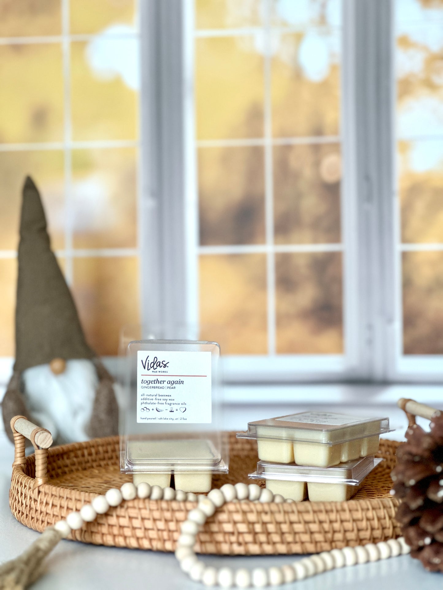 Embrace autumn's enchantment: Gingerbread and pear fragrance. An open 2.5oz clamshell sits on top of a wicker serving tray beside two stacked clamshell packages. A string of beads and a pinecone adorn the foreground, while a charming brown gnome lends whimsy to the scene against the backdrop of blurred yellow fall leaves outside a window.