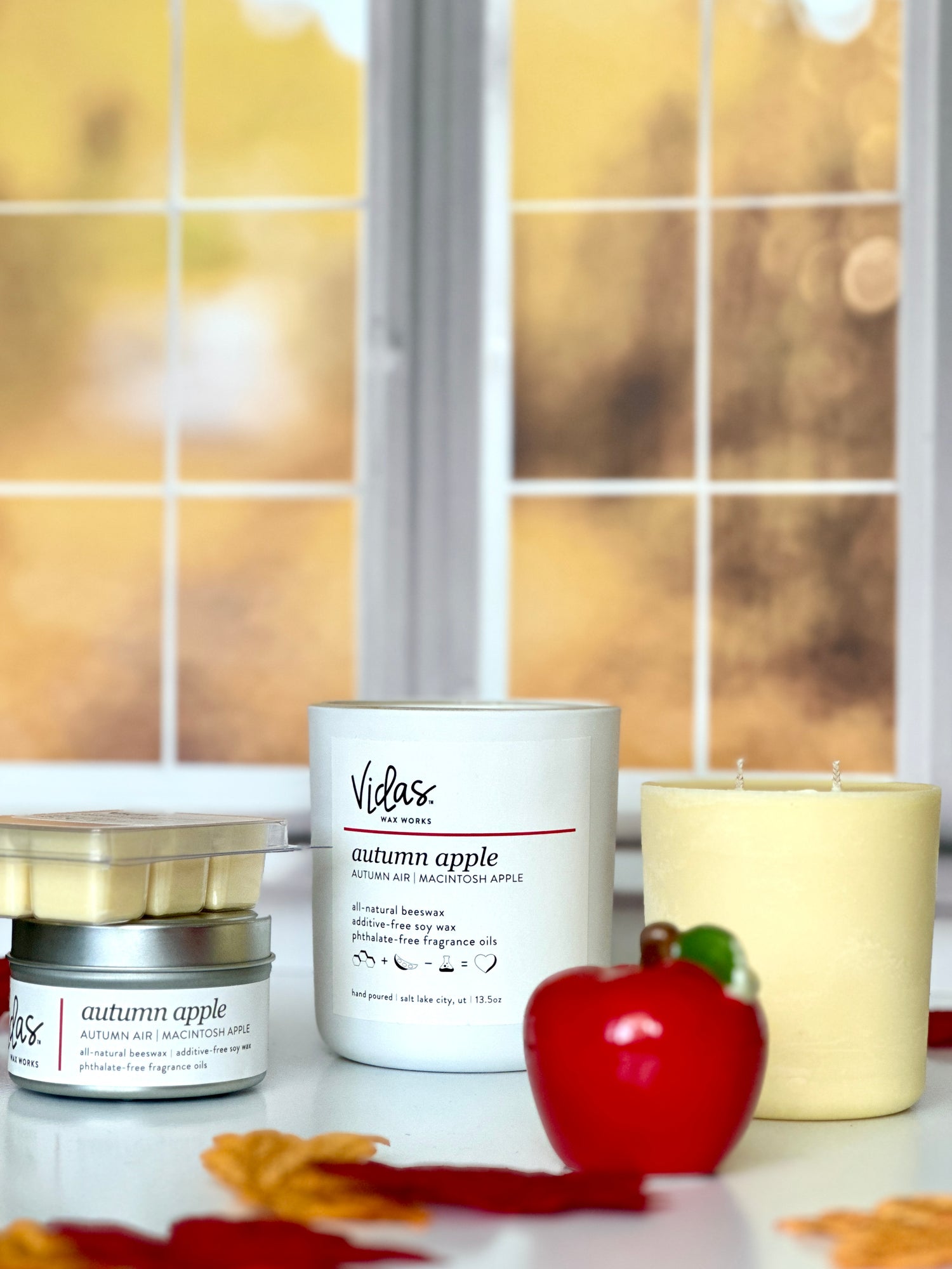 Embrace the autumn ambience with our “Autumn Apple” scent, a delightful blend of autumn air and macintosh apple. Against a backdrop of blurred fall-colored leaves outside a window, the collection features a range of offerings: a 13.5oz candle in a chic matte white jar, a 13.5oz candle refill, a compact 3.5oz candle tin, and dainty 2.5oz wax melts. A symphony of scents to elevate your seasonal moments.