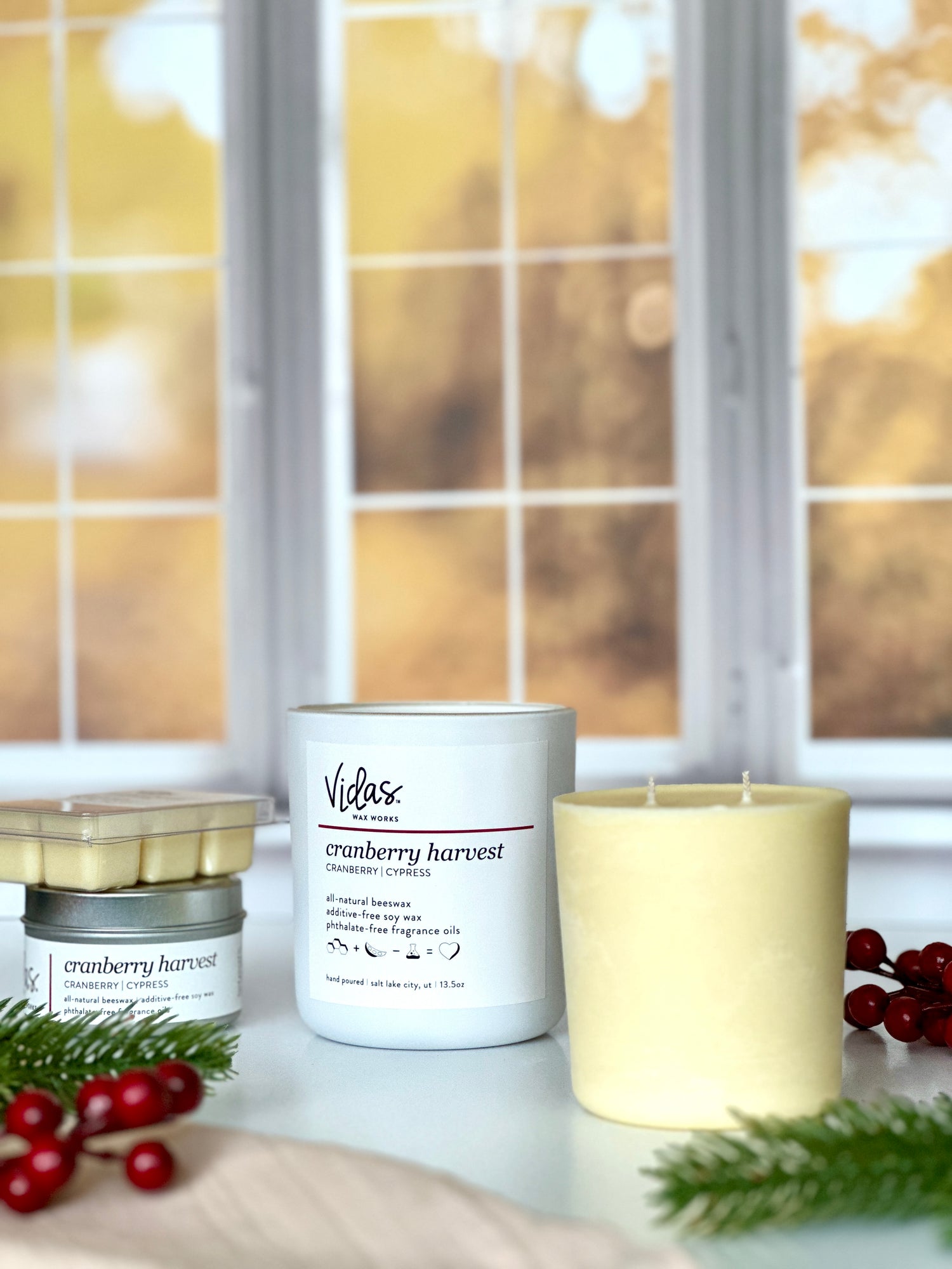 Embrace the autumn ambience with our “Cranberry Harvest” scent, a delightful blend of cranberry and cypress. Against a backdrop of blurred fall-colored leaves outside a window, the collection features a range of offerings: a 13.5oz candle in a chic matte white jar, a 13.5oz candle refill, a compact 3.5oz candle tin, and dainty 2.5oz wax melts. A symphony of scents to elevate your seasonal moments.