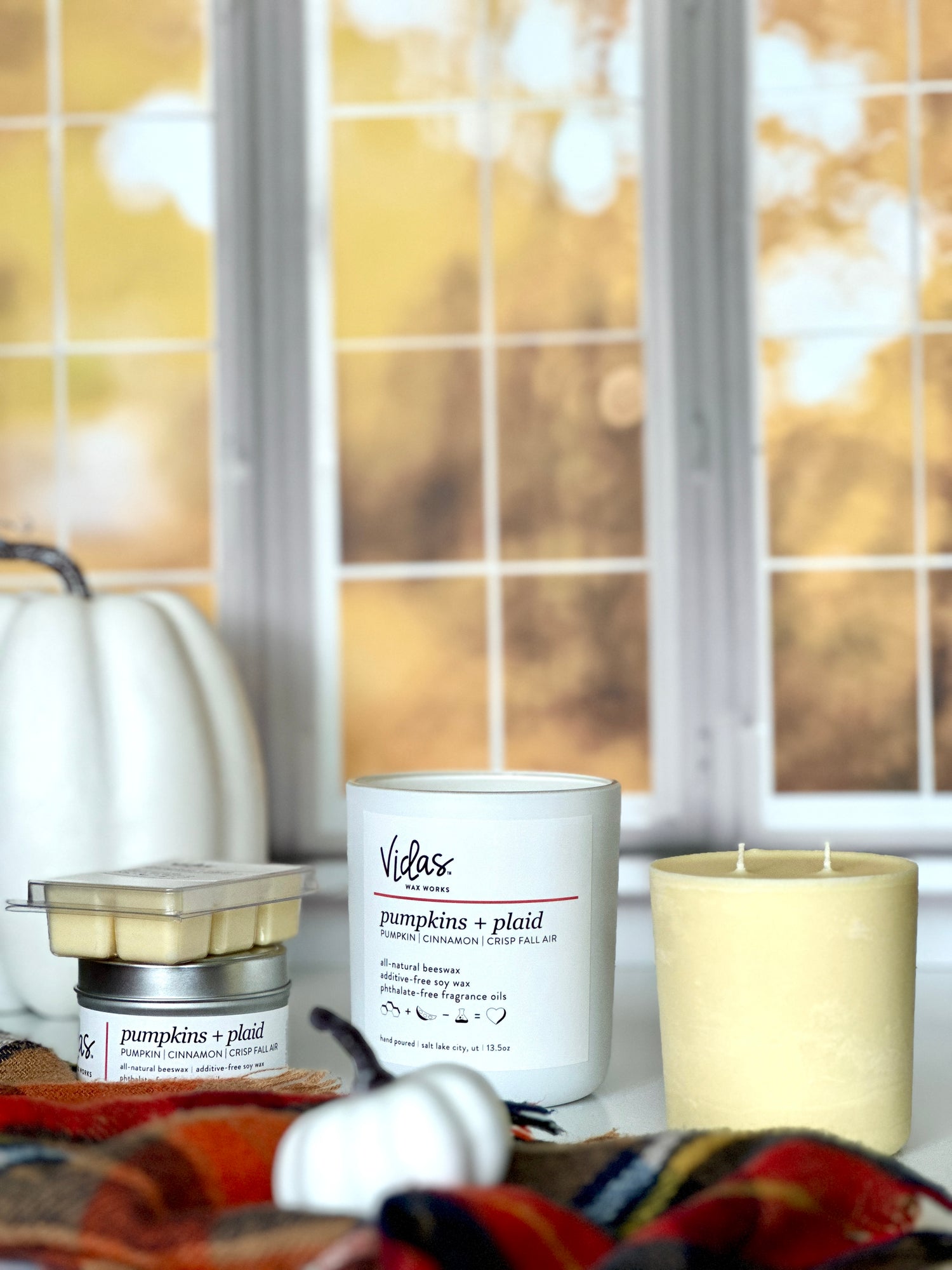Embrace the autumn ambience with our "Pumpkins + Plaid" scent, a delightful blend of pumpkin, cinnamon, and crisp fall air. Against a backdrop of blurred fall-colored leaves outside a window, the collection features a range of offerings: a 13.5oz candle in a chic matte white jar, a 13.5oz candle refill, a compact 3.5oz candle tin, and dainty 2.5oz wax melts. A symphony of scents to elevate your seasonal moments.